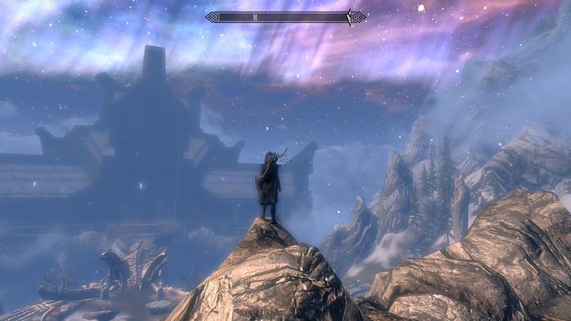 Screenshot from the game Skyrim with character on a high rock overlooking a night sky and a building in the distance