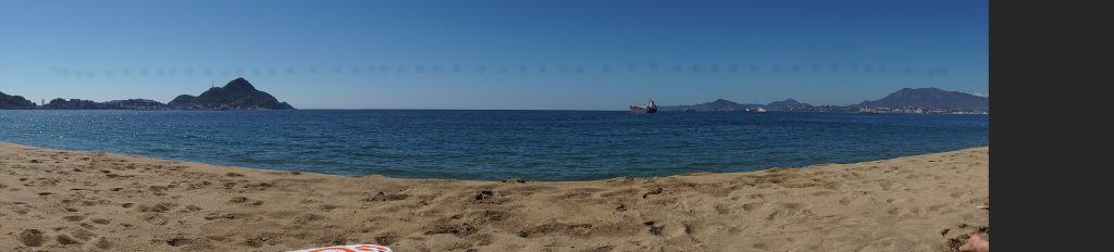 Panoramic shot at Las Brisas beach in Manzanillo. Sand in the foreground with a view of the bay, some ships visible.