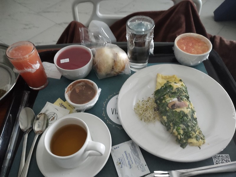 Tray of breakfast in the hospital: omelette, tea, juice, fruit, jello, break, glass of water.