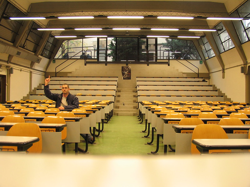 Lone person in a desk of an otherwise empty classroom raises his hand to ask a question.