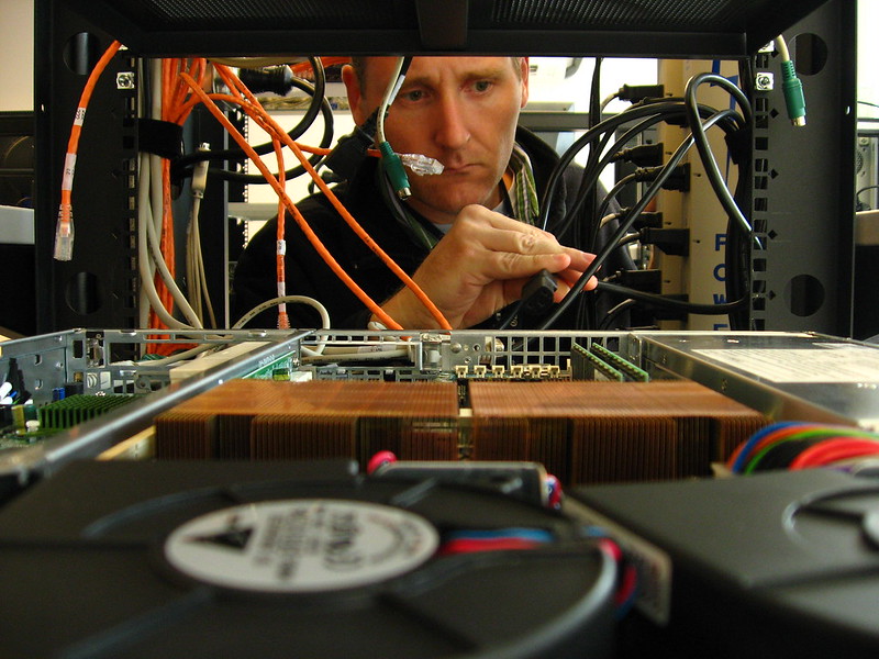 Tight shot of a man framed by a server rack with a open rackmount computer in the foreground. He is plugging in a power supply cable.