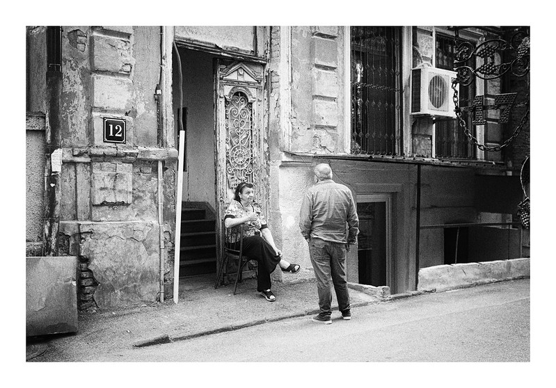 A black and white street image of a woman seated just outside a doorway talking to a man with his back to us standing on the edge of the road. Older building in the background. Apparently the shot is taken at Tbilissi, Georgia.