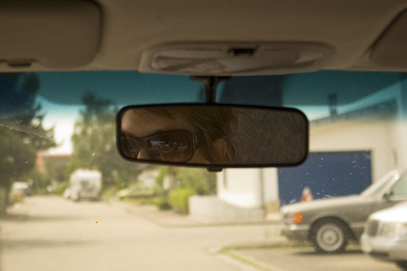Close shot from back seat of a car with a view of the drivers glasses via the rear-view mirror of the vehicle. Residencial street (out of focus from shallow depth of field) visible through the windshield.