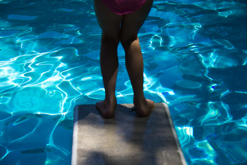 Close up of legs and pink bathing trunks about to jump from a standard height diving board into the blue of a swimming pool which takes up the rest of the image.