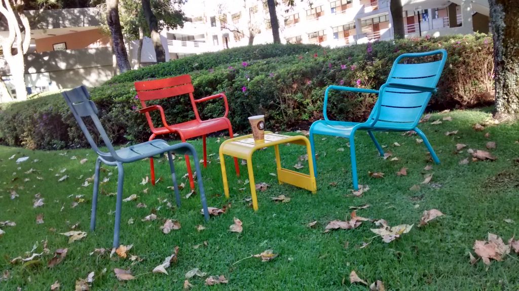 Three coloured (grey, orange, blue) metal lawn chairs arranged around a similar style yellow small table with a coffee on it. This on the grass in the gardens of the Tecnológico de Monterrey in Guadalajara.