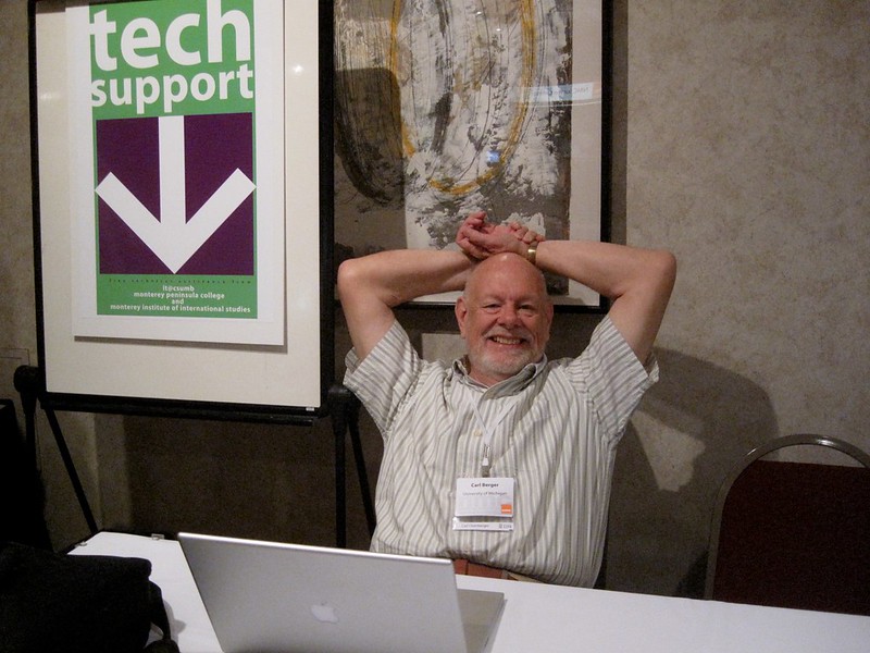 Carl Berger sitting behind a conference event table in front of an Apple laptop with a "Tech Support" sign behind and to his right.