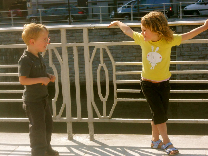Two young children speaking to each other about 1 meter apart near a fence, one is leaning back against the fence with her arms up and out.