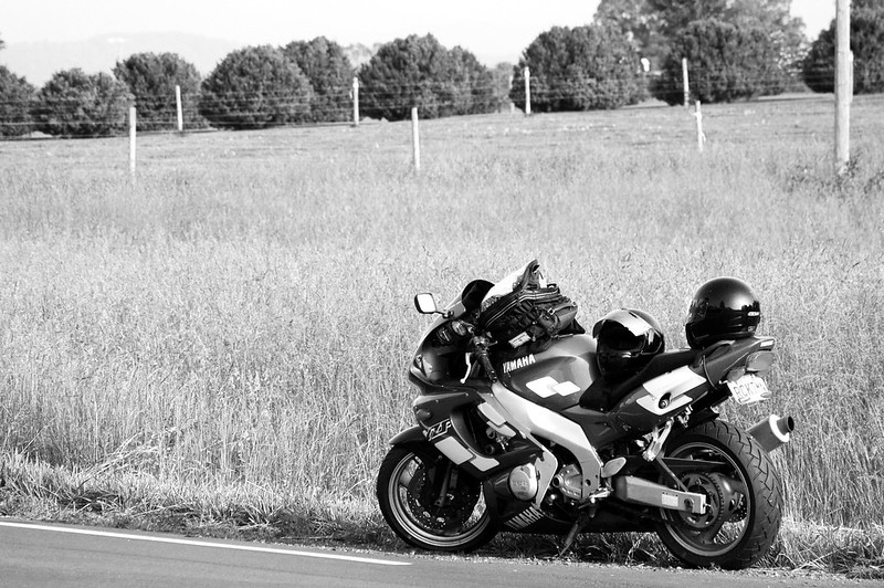 Black and white picture of a Yamaha sport motorcyle on the side of the road with long grass, fence and trees in the background.
