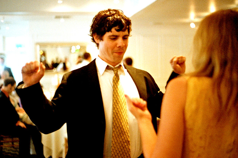 View of a couple dancing at a wedding. Women with her back to us; man in suit and tie with arms ready to pump up while busting out a dance move.