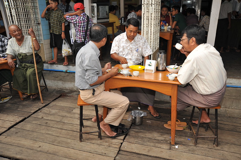 A tea house in Mandalay, Myanmar. Various people in the background inside the tea house with three men at a short table in the foreground drinking tea together on a wooden deck outside the entrance.